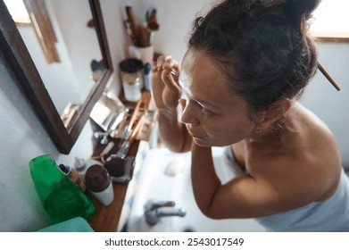 Woman focusing on her makeup in a bathroom mirror, surrounded by various cosmetics. Morning routine emphasizes beauty and grooming, reflecting a sense of care and personal preparation. - Powered by Shutterstock