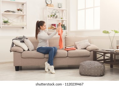Woman folding clean t-shirts, sitting on sofa at home. Young girl tidying up clothes after laundry or shopping, copy space