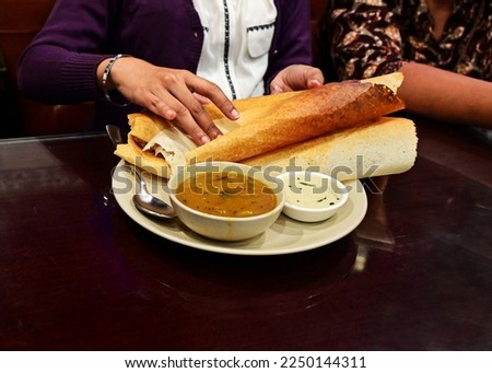 Woman folding a rolled two-foot Ghee roast Dosa, a thin, crispy South Indian dish made of rice and black gram roasted after smearing with clarified butter, in a restaurant. Sambar and chutney.