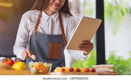 A Woman Following Recipe On Digital Tablet While Cooking Salad And Sandwich In The Kitchen, Online Learning Cooking Class Concept