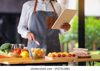 A Woman Following Recipe On Digital Tablet While Cooking Salad And Sandwich In The Kitchen, Online Learning Cooking Class Concept