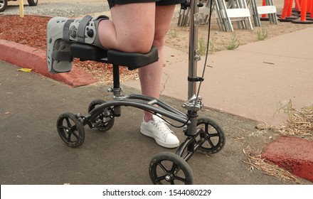 Woman With Foot Injury Getting Around With The Help Of A Walking Cast And A Knee Scooter. 