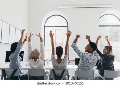 Woman In Formal Shirt With Blonde Hair Waving Hands, Sitting Between Coworkers In Big Light Conference Hall. Photo From Back Of Tired Managers Stretching During Meeting In Office.