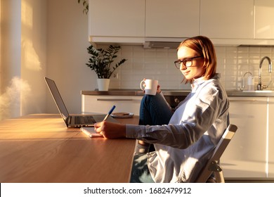 Woman freelancer in glasses using laptop and makes notes in a notebook, works from home during quarantine due to coronavirus, hold coffee/tea mug. Cozy office workplace, remote working, stay home 