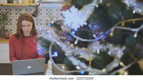 Woman freelancer working remotely on christmas at laptop, portrait. Decorated Christmas tree flashing garland. Indoors, living room kitchen. - Shutterstock ID 2255723337