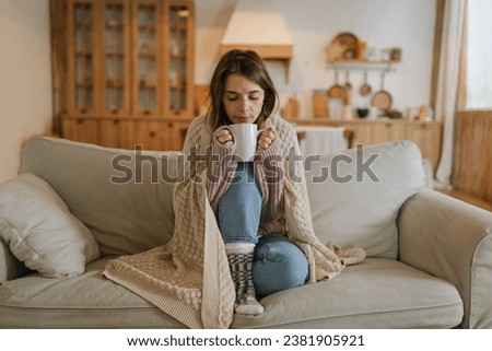 Woman freezes in winter time. Young girl wearing warm woolen socks and wrapped into plaid, holding a cup of hot drink while sitting on sofa at home. Keep warm. 