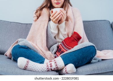 Woman freezes in wintertime. Young girl wearing warm woolen socks and wrapped into two blankets, holding a cup of hot drink and heating pad while sitting on sofa at home. Keep warm. Selective focus
