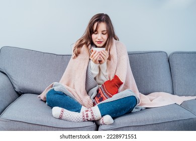 Woman freezes in wintertime. Young girl wearing warm woolen socks and wrapped into two blankets, holding a cup of hot drink and heating pad while sitting on sofa at home. Keep warm. Selective focus. - Shutterstock ID 2228971551