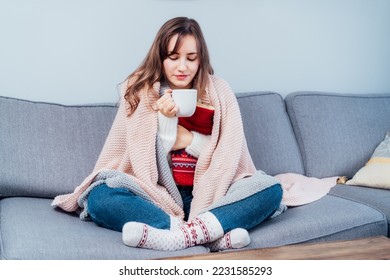 Woman freezes in wintertime. Young girl wearing warm woolen socks and wrapped into two blankets, holding a cup of hot drink and heating pad while sitting on sofa at home. Keep warm. Selective focus - Shutterstock ID 2231585293
