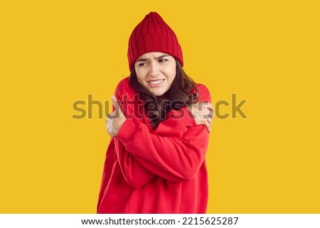 Woman freezing in cold autumn or winter weather. Young brunette girl wearing warm wool red hat and sweatshirt standing on yellow background, feeling very cold and shivering with sad face expression