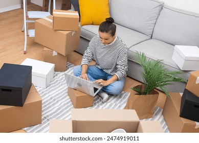 Woman in freshly acquired apartment, amidst boxes, works on her laptop, exploring interior design, online shopping, or arranging moving services - Powered by Shutterstock