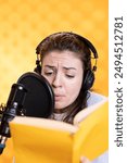 Woman frowns while doing voiceover reading of book to produce audiobook using melodramatic acting. Narrator glowers, using storytelling skills to portray character, recording novel, studio background