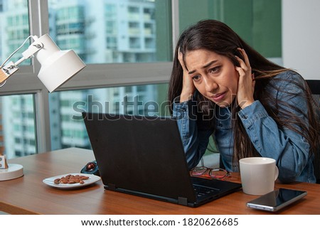 Woman frustrated and tired looking at the laptop, holding her head with her hands in a wooden desk. With a cup of coffee and a cell phone next to her
