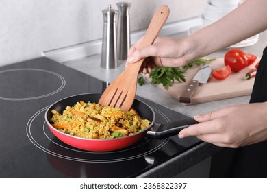 Woman frying rice with meat and vegetables on induction stove in kitchen, closeup