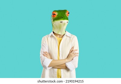 Woman In Funny Animal Disguise. Portrait Of Strange Lady Wearing Silly Eccentric Green Frog Mask With Thoughtful Snobby Sceptical Face Expression Standing With Her Arms Folded On Turquoise