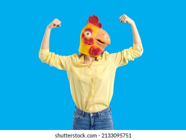 Woman in funny chicken disguise pretends to be super strong. Portrait of confident young girl wearing strange weird ridiculous absurd bizarre Hallowen mask flexing her arms isolated on blue background