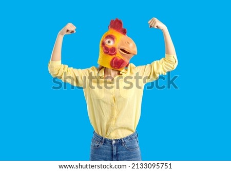Woman in funny chicken disguise pretends to be super strong. Portrait of confident young girl wearing strange weird ridiculous absurd bizarre Hallowen mask flexing her arms isolated on blue background