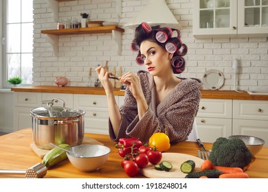 Woman In Funny Curlers Sitting At Kitchen Table And Filing Nails With Thoughtful Face Expression. Angry Wife Waiting For Husband From Work. Young Housewife Cooking, Thinking And Making Cunning Plans