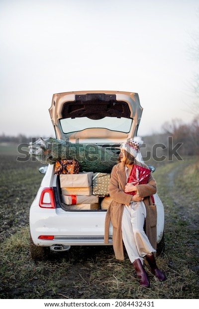 Woman in fur\
coat and winter hat sitting at car trunk full of giftboxes and\
Christmas tree on the field at sunset. Concept of New Year\'s mood.\
Idea of preparation for the\
holiday