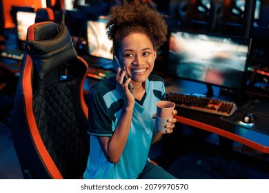 Woman Gamer Sitting On Streaming Chair With Coffee Cup And Sharing Positive Emotions With Friend By Phone In Cyber Club