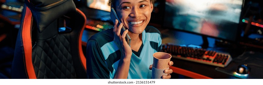 Woman Gamer Sitting On Streaming Chair With Coffee Cup And Sharing Positive Emotions With Friend By Phone In Cyber Club