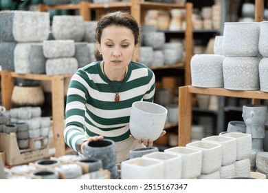 Woman in garden goods store carefully chooses flower pot, studies assortment, admires variations ceramic products. Concept of self-decoration of housing, indoor plants - Powered by Shutterstock