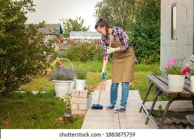 Woman Gardener In An Apron And Gloves Sweeps The Path With A Broom In Her Garden. A Young Girl Takes Care Of Her Dacha Plot. Working In Her Garden Near House.