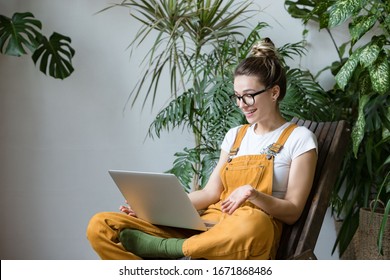 Woman Gardener In Glasses Wearing Overalls, Sitting On Wooden Chair In Greenhouse, Using Laptop After Work, Talks With Her Friend About Coronavirus And Stay Home During Online Video Call. Working Home