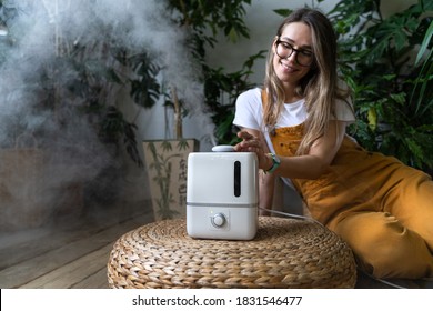 Woman gardener in overalls feeling fresh, sitting on the wooden floor, using air humidifier in home garden during heating period, houseplants on background. Plant care. 