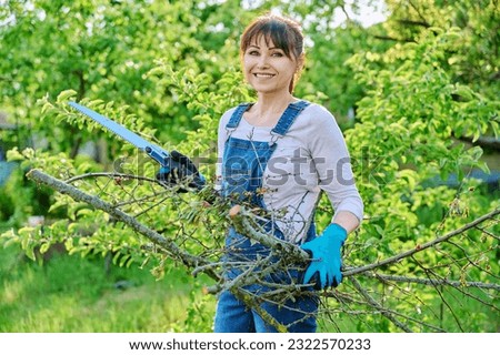 Woman gardener with saw and dry cut branches looking at camera in garden