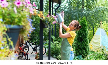 A woman gardener waters flowers in a hanging planter. A female farmer in a green apron tending a blooming garden on a sunny summer day. Ideas for vertical gardening of landscape design. - Powered by Shutterstock