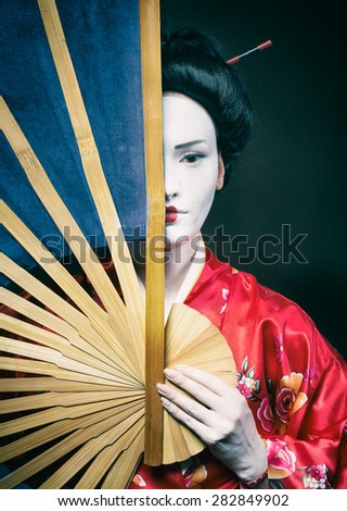 Woman in geisha makeup covering half of her face with a big fan