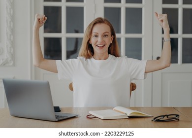 Woman Gestures Power. Young Tender Businesswoman Is Working At Laptop. Happy European Lady In White T-shirt Is Freelancer Having Remote Work In Her Apartment. Concept Of Hard Work And Success.