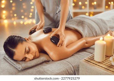 Woman getting exotic spa massage with hot stones. Happy, relaxed young woman lying on spa bed while professional masseuse is putting hot stones on her back. Spa treatment, body relaxation concept - Powered by Shutterstock