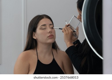 A woman is getting her makeup done by a woman in a black shirt. The woman in the black shirt is using a brush to apply makeup to the woman's face - Powered by Shutterstock