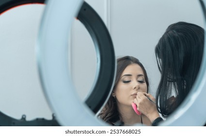 A woman is getting her makeup done by another woman. The makeup artist is using a pink brush to apply makeup to the woman's face. The woman is looking at herself in the mirror - Powered by Shutterstock