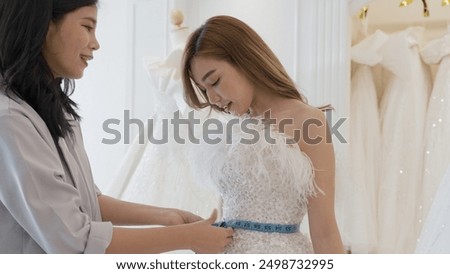 A woman is getting her wedding dress measured by a tailor. The tailor is holding a blue tape measure and the woman is smiling.