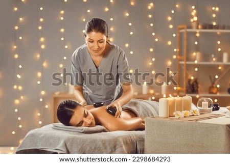Woman getting hot stones massage at massage salon or spa room with lights and candles. Relaxed young woman lying on spa bed while professional masseuse puts hot stones on her back