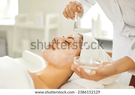 Woman getting professional skin facial care treatment procedures at spa and beauty salon. Young lady with white towel turban on head lying on bed while beautician applies cleansing mask on her face