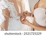 A woman is getting ready for a wedding and her friend is helping her with her dress. The bride is wearing a white dress with lace detailing and a veil. The bride