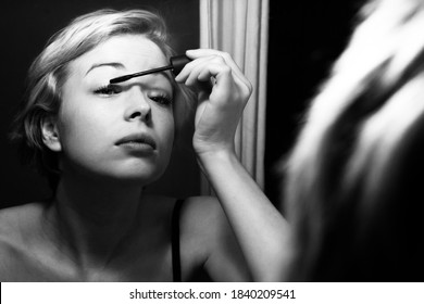 Woman Getting Ready For Work Doing Morning Makeup Routine Applying Mascara In Bathroom Mirror At Home. Beautiful Caucasian Girl Applying Eye Make-up. Black And White Image.