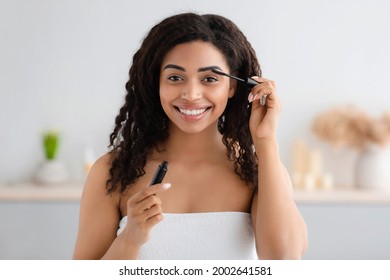 Woman Getting Ready For Work, Doing Morning Makeup Routine In Bathroom At Home. Glad Millennial Black Woman In White Towel Applies Mascara With Brush On Eyelashes In Bright Interior, Empty Space