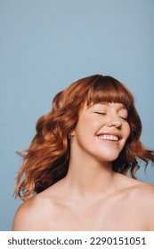 Woman with ginger hair and flawless skin smiling happily with her eyes closed. Confident young woman feeling comfortable in her own skin. Young woman standing topless against a blue studio background. - Shutterstock ID 2290151051