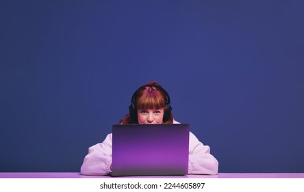 Woman with ginger hair looking at the camera while sitting behind her gaming laptop in a studio. Female gamer using an interactive streaming service while sitting in neon purple light. - Shutterstock ID 2244605897
