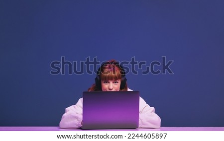 Woman with ginger hair looking at the camera while sitting behind her gaming laptop in a studio. Female gamer using an interactive streaming service while sitting in neon purple light.