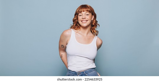 Woman with ginger hair smiling at the camera while wearing a tank top and jeans. Portrait of a happy young woman standing against a blue background. - Powered by Shutterstock