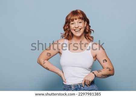 Woman with ginger hair smiling at the camera in a studio. Portrait of a happy young woman standing in a white tank top and jeans.