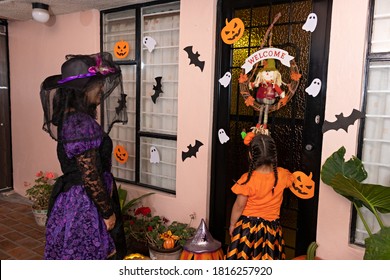 A Woman And A Girl In Disguise Are Going To Knock On The Door Of A House With Halloween Decoration