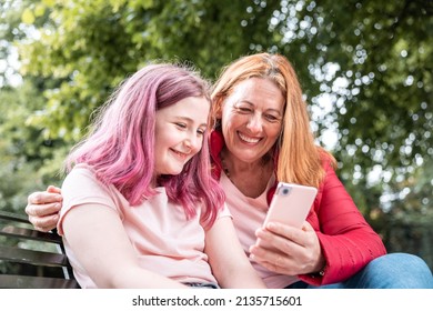 Woman And Girl Looking At Mobile Phone At Park And Smiling - Mother And Daughter Having Fun Together Sharing On Social Media With Smartphone - Summer And Lifestyle Concepts