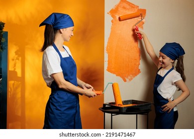 Woman And Girl Painting Room Walls With Orange Color Paint To Renovate Apartment Interior With Tools. Small Family Redecorating House Room With Paintbrush Roller, Home Renovation.